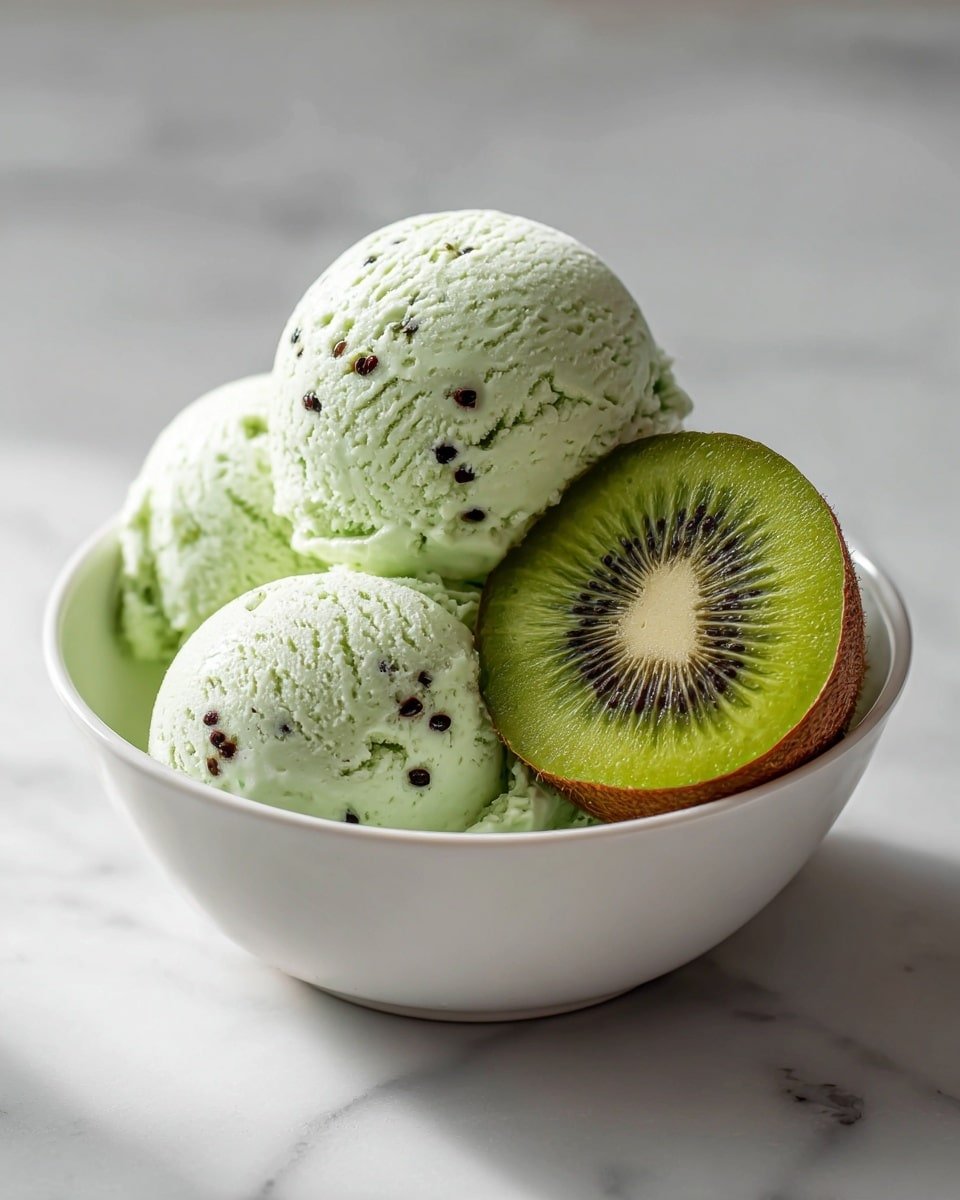 A white bowl filled with three scoops of light green ice cream with tiny black seeds scattered on the surface, showing a rough, frosty texture. Behind the scoops, a slice of fresh green kiwi with dark seeds and a brown edge leans inside the bowl. The white bowl sits on a white marbled surface, and in the background, blurry whole and halved kiwis add a fresh look to the scene. Photo taken with an iphone --ar 4:5 --v 7 — Kiwi Sorbet, refreshing kiwi dessert, homemade sorbet, healthy summer treat, easy fruit sorbet