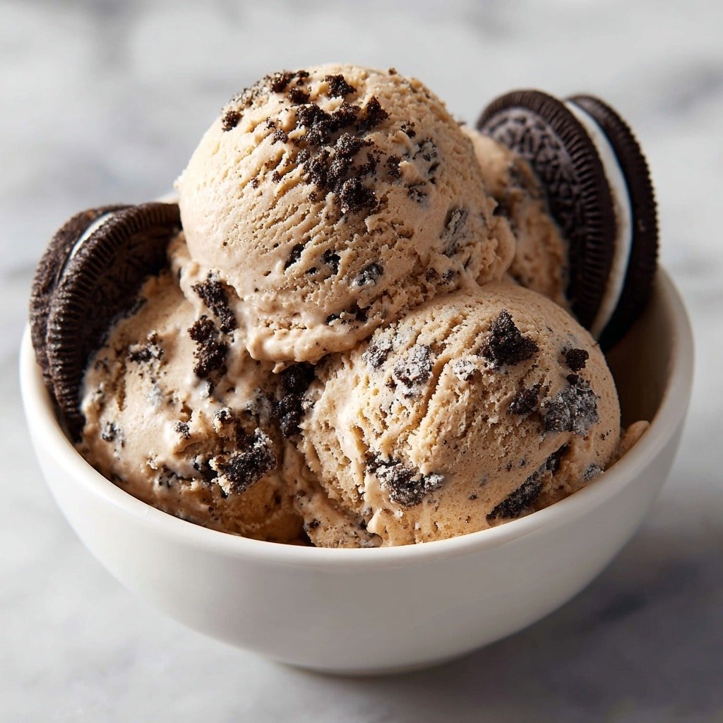 A close-up view showing three scoops of light brown ice cream with dark cookie pieces mixed inside, placed closely together in a white bowl. The ice cream texture looks creamy and slightly soft with some crumbly dark cookie bits scattered on the surface. Two whole chocolate sandwich cookies with white cream filling are partially pressed into the ice cream on the right and left sides, adding a crunchy contrast. The bowl rests on a white marbled surface. photo taken with an iphone --ar 1:1 --v 7 — Chocolate Oreo Protein Ice Cream, healthy ice cream recipe, homemade protein dessert, low-calorie ice cream, Oreo protein ice cream