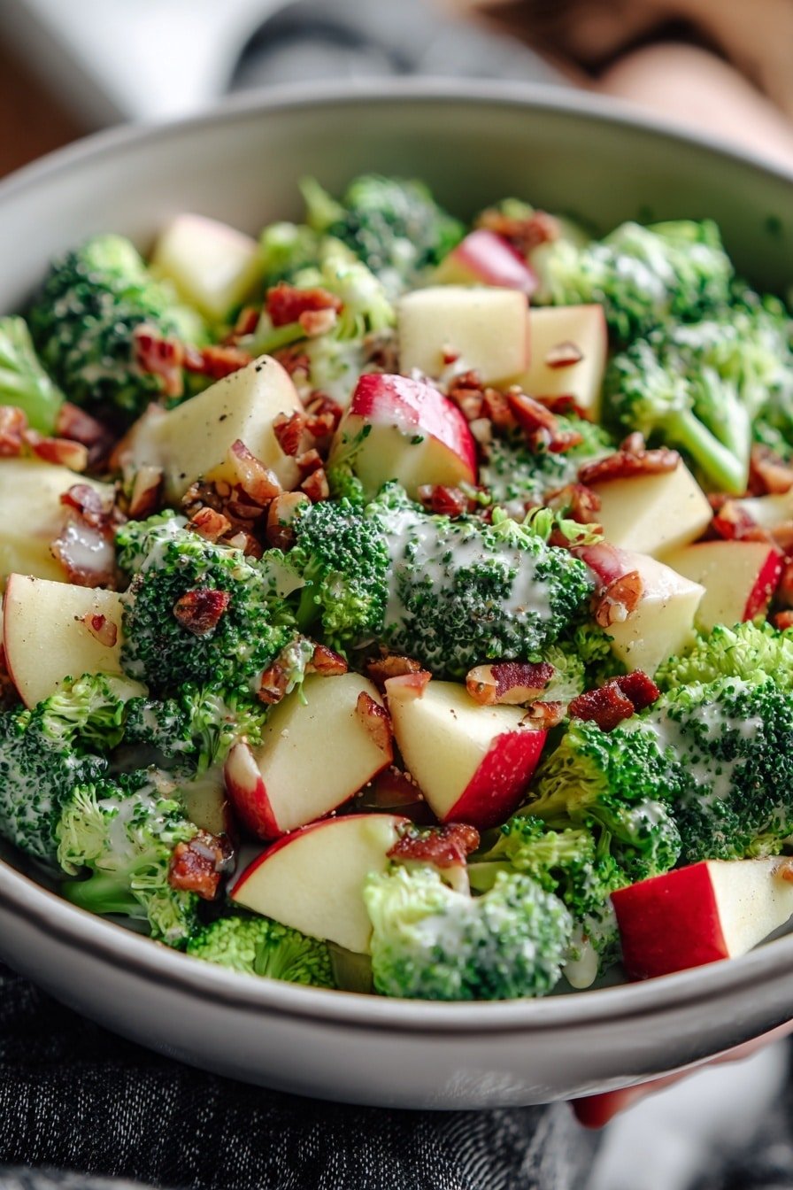 The image shows a close-up of a white bowl filled with a fresh salad on a white marbled surface. The salad has three clear layers: a bottom layer of bright green broccoli florets with a bumpy texture, a middle layer of red apple chunks with shiny reddish skin and pale inside, and a top layer of small brown chopped nuts to add crunch. A creamy light dressing is lightly drizzled over the salad, giving a slight shine on some broccoli and apple pieces. In the background, a woman's hand is slightly visible holding the bowl. Photo taken with an iphone --ar 2:3 --v 7 — Honeycrisp Apple Broccoli Salad, healthy broccoli apple salad, easy fruit and vegetable salad, tangy honey mayo dressing, refreshing apple broccoli side dish