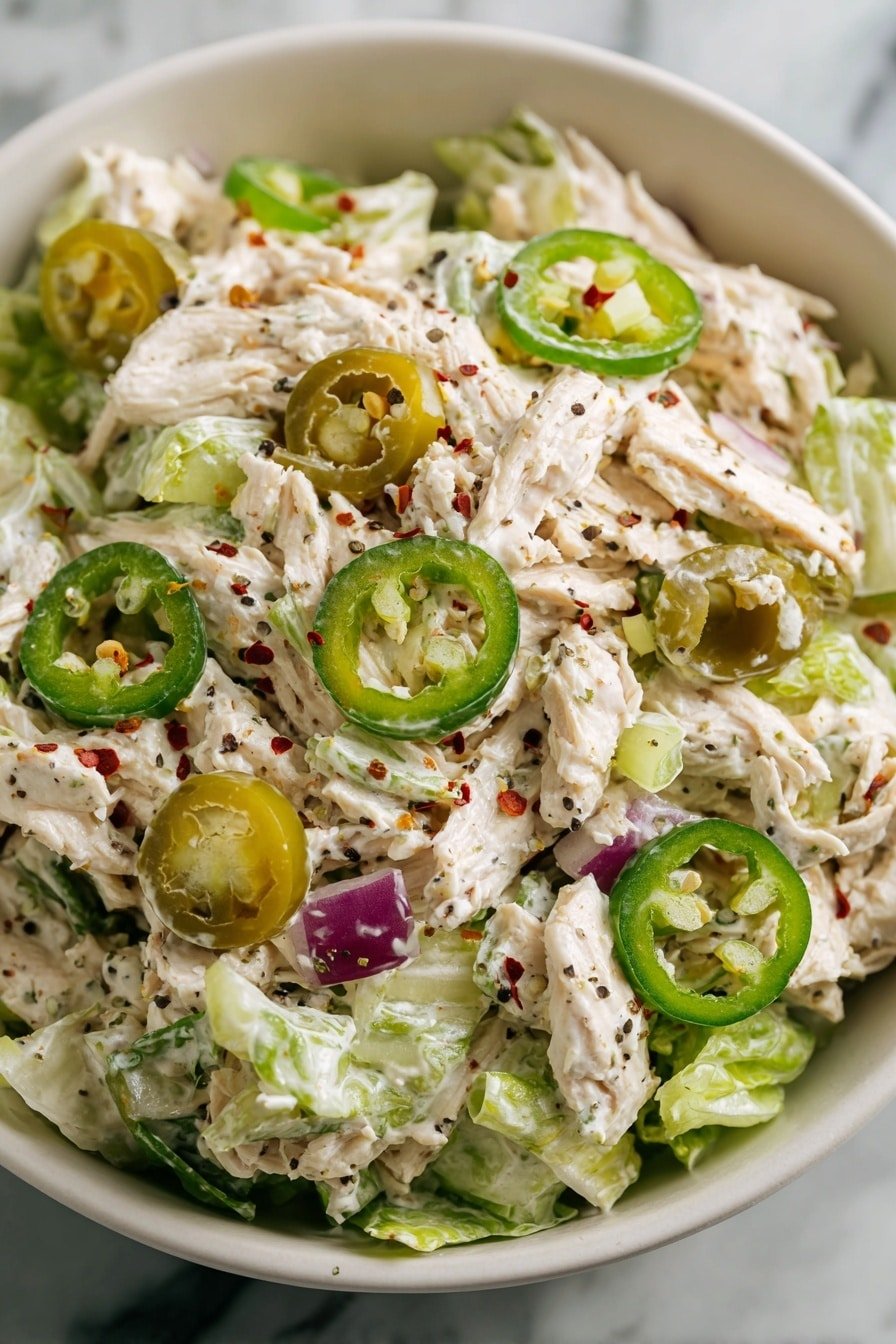 The dish is a creamy chicken salad served in a white bowl placed on a white marbled surface. The salad has three visible layers: the bottom layer consists of light green lettuce pieces, the middle layer is filled with shredded white chicken mixed with creamy dressing along with chopped celery and purple onion bits, and the top layer is decorated with green jalapeño rings scattered over the salad. The creamy dressing lightly coats the chicken and vegetables, with a sprinkle of black pepper and red chili flakes adding specks of red and black texture throughout the dish. photo taken with an iphone --ar 2:3 --v 7 — High Protein Pepperoncini Chicken Salad, healthy chicken salad recipes, high-protein lunch ideas, easy chicken salad with Greek yogurt, flavorful protein salads