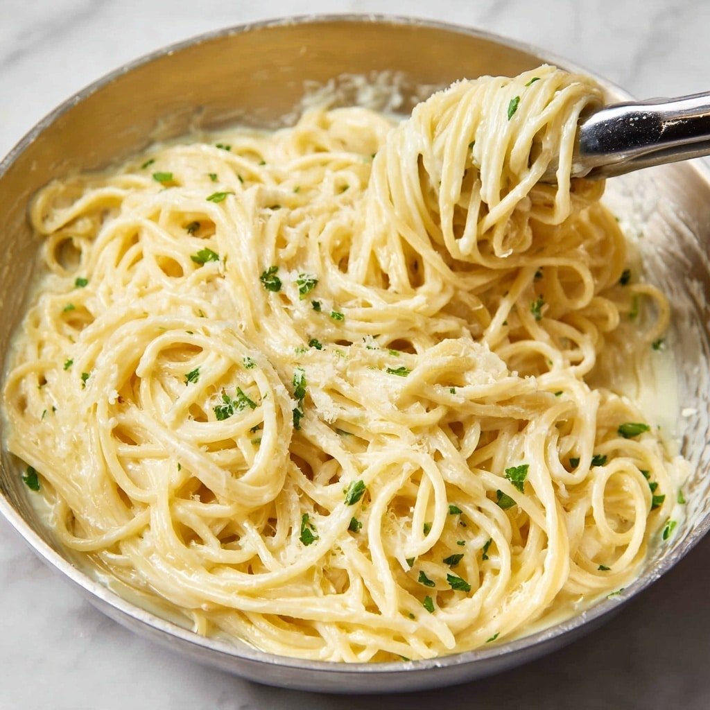 A close-up view of a large white metal bowl filled with creamy spaghetti noodles, coated in a smooth, pale yellow sauce. The pasta is twisted and piled high, with small green herb pieces sprinkled evenly on top. A pair of silver tongs hold a bunch of noodles in the upper right, lifting them slightly above the rest. The bowl sits on a white marbled surface. Photo taken with an iphone --ar 1:1 --v 7 — Garlic Parmesan Pasta, Garlic Parmesan Pasta Recipe, Creamy Garlic Pasta, Easy Pasta Dinner, Quick Pasta Recipes